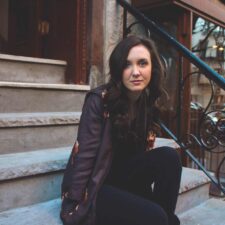 a young white woman in black clothing and a brown leather jacket sits on a concrete staircase. she has brown hair slightly past her shoulders and is looking intently at the camera.