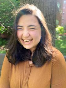 a young woman smiles into the camera, clearly laughing. she is mixed race--Japanese and Jewish-- and wears a bronze shirt. her dark brown hair rests past her shoulders, and there are trees in the background