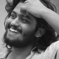 a black and white photo of a brown-skinned man. he smiles and looks away from the camera, off into the distance, with his hand on his forehead. he has dark hair on his head and face.