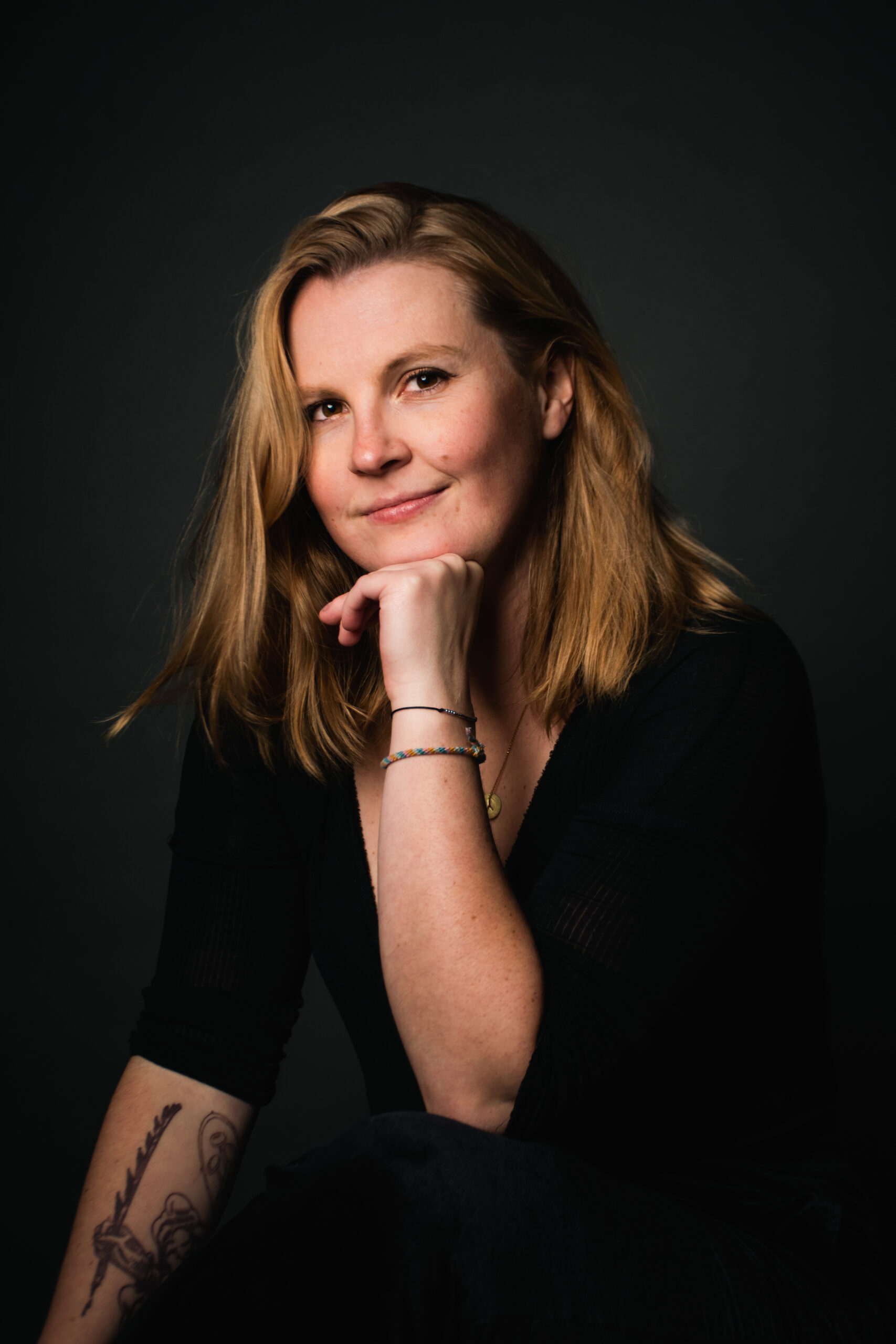 Emily Breeze, a white playwright with long blonde hair, rests her chin on her left hand while looking into the camera. She has a mischievous smile and a tattoo along her right arm. She's wearing a bracelet on her left arm, a black shirt and is against a black background.