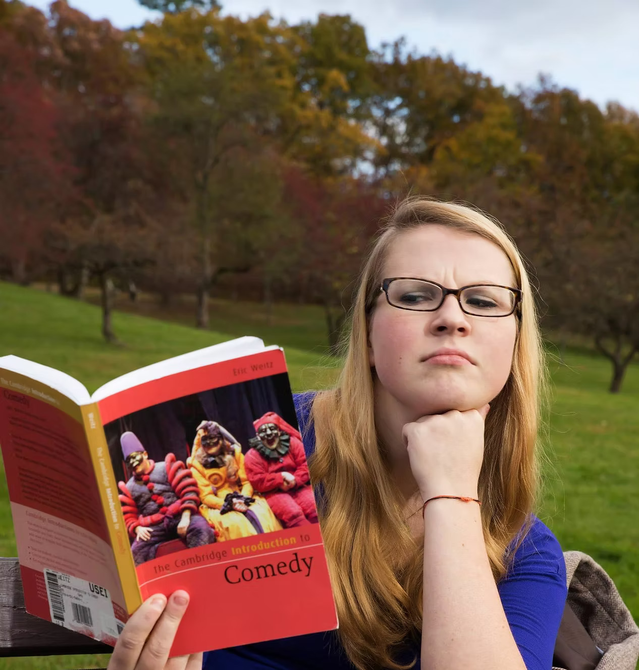 Emily Breeze, a white playwright with long blonde hair and a deep blue shirt, reads with a quizzical look from her red book, titled "Comedy." She squints through her glasses at the book while resting her hand on her chin. She is sitting in a grassy area with trees in the background.