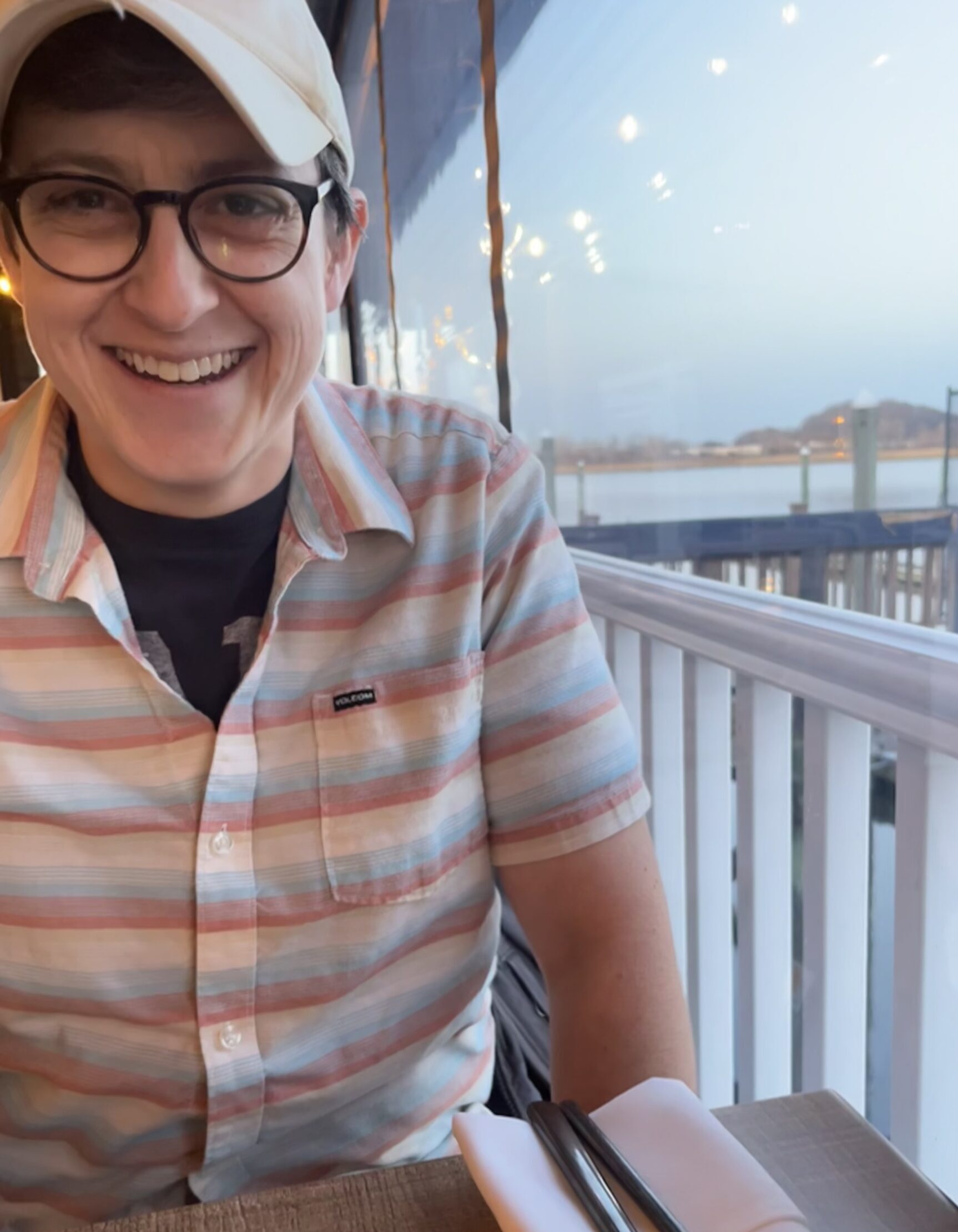 Katie Chance, a white woman with thick framed glasses and a tan baseball cap, smiles at the camera. She is wearing a button up shirt with light blue, pink, and white stripes. She is seated at a wooden table. A window is to her right, overlooking a body of water.