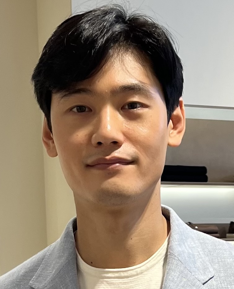 Matthew Chong, a Korean-American man, looks into the camera with a slight smile. His black hair is swept to his right. He is wearing a grayish-blue jacket over a white shirt.