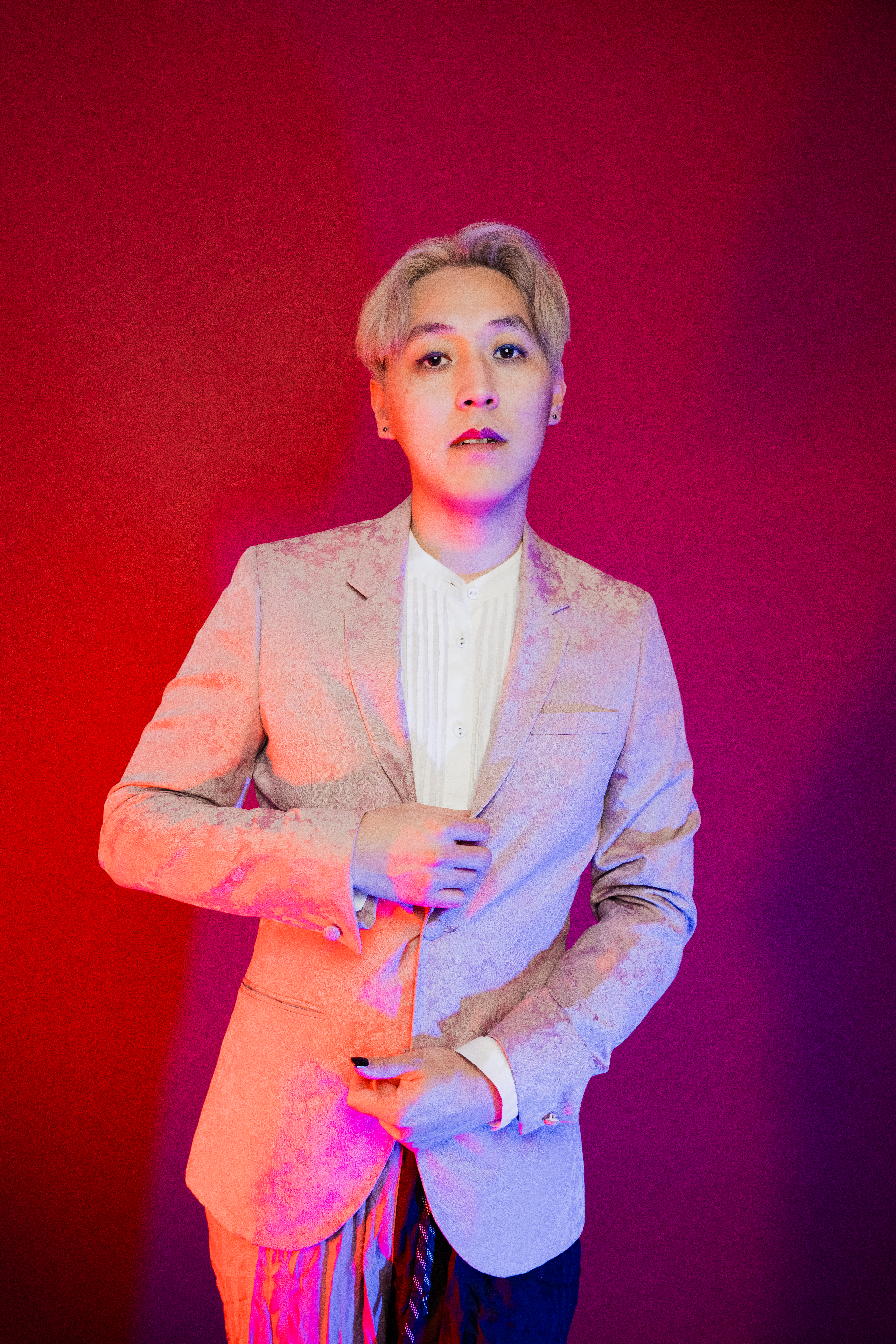 Gavin D. Pak, a Korean American person, looks into the camera. They have bleach blonde hair and lipstick on their upper lip. They are wearing a white button up and a rose print, pink suit. Their right hand adjusts their button. They are against a red background and are lit with red lights on their right side and blue lights on their left.