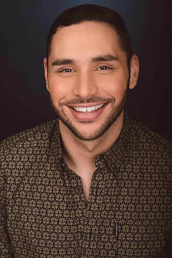Andrew Rincón, a Colombian-American playwright, smiles at the camera with a relaxed look. His hair is cut short, and he sports a well-kept, short beard. He is wearing a patterned brown button-up shirt.