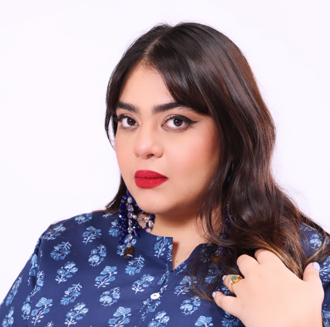 Sarah Saifi, a Pakistani woman, angles her head to her right and looks into the camera. She has dark, long hair, winged eyeliner, and red lipstick. Her left hand, with a ring on her ring finger, touches her own shoulder. She is wearing a blue blouse with white floral patterning.