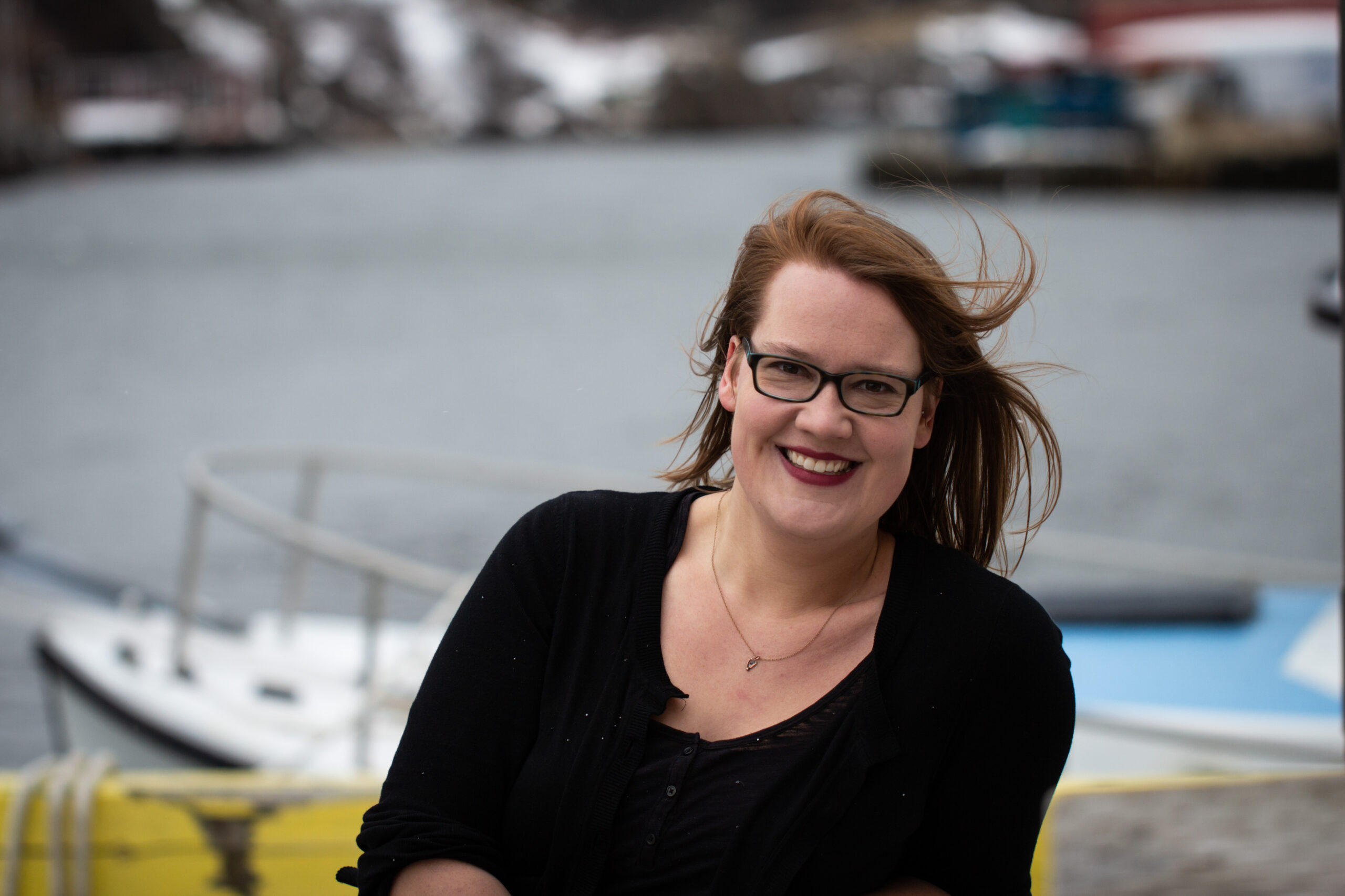 Mara is a white person with shoulder-length reddish-brown hair and thick-framed glasses. She is seen from the waist up wearing a black shirt and cardigan. She is smiling at the camera because it is definitely not freezing cold in the harbour.